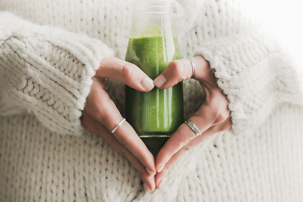 Hände formen ein Herz um ein Glas mit grünem Smoothie. Das Bild symbolisiert die herzliche Betreuung bei der enteralen Ernährungstherapie.